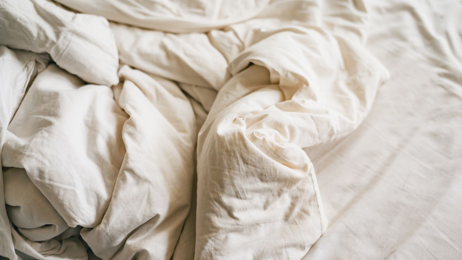 Crumpled oatmeal linen duvet and sheets on a bed in soft morning light, showing the natural wrinkle of washed flax